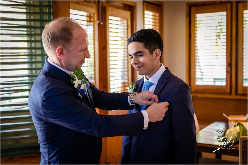 Two people in formal attire share a moment while adjusting boutonnieres before a ceremony.