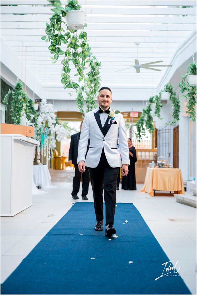 A groom walks down a blue carpet aisle in a light gray tuxedo jacket inside an elegant venue with hanging greenery.