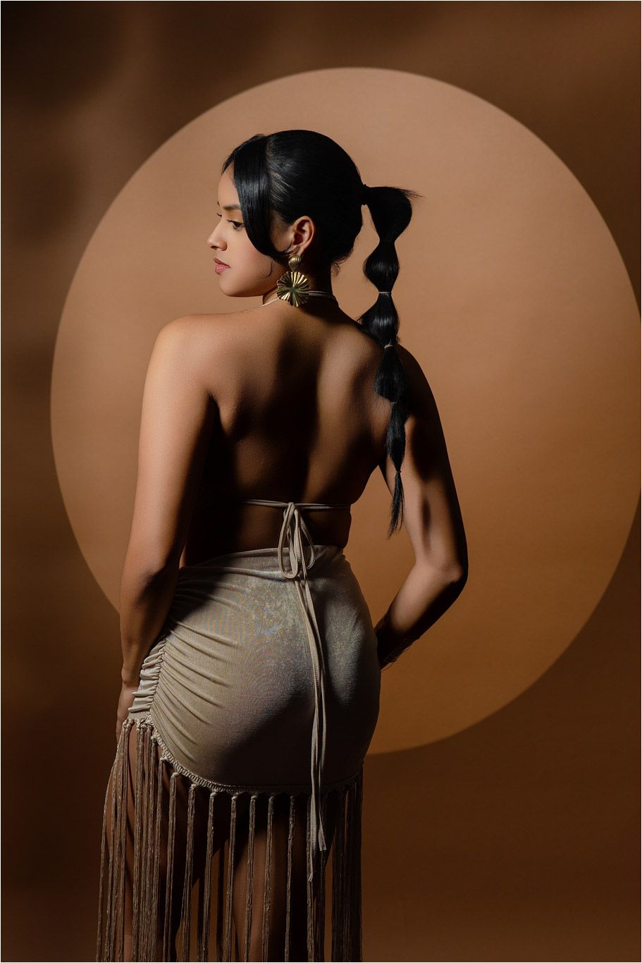 Woman in backless tan fringe skirt with bubble braid hairstyle poses against warm brown background with circle detail.