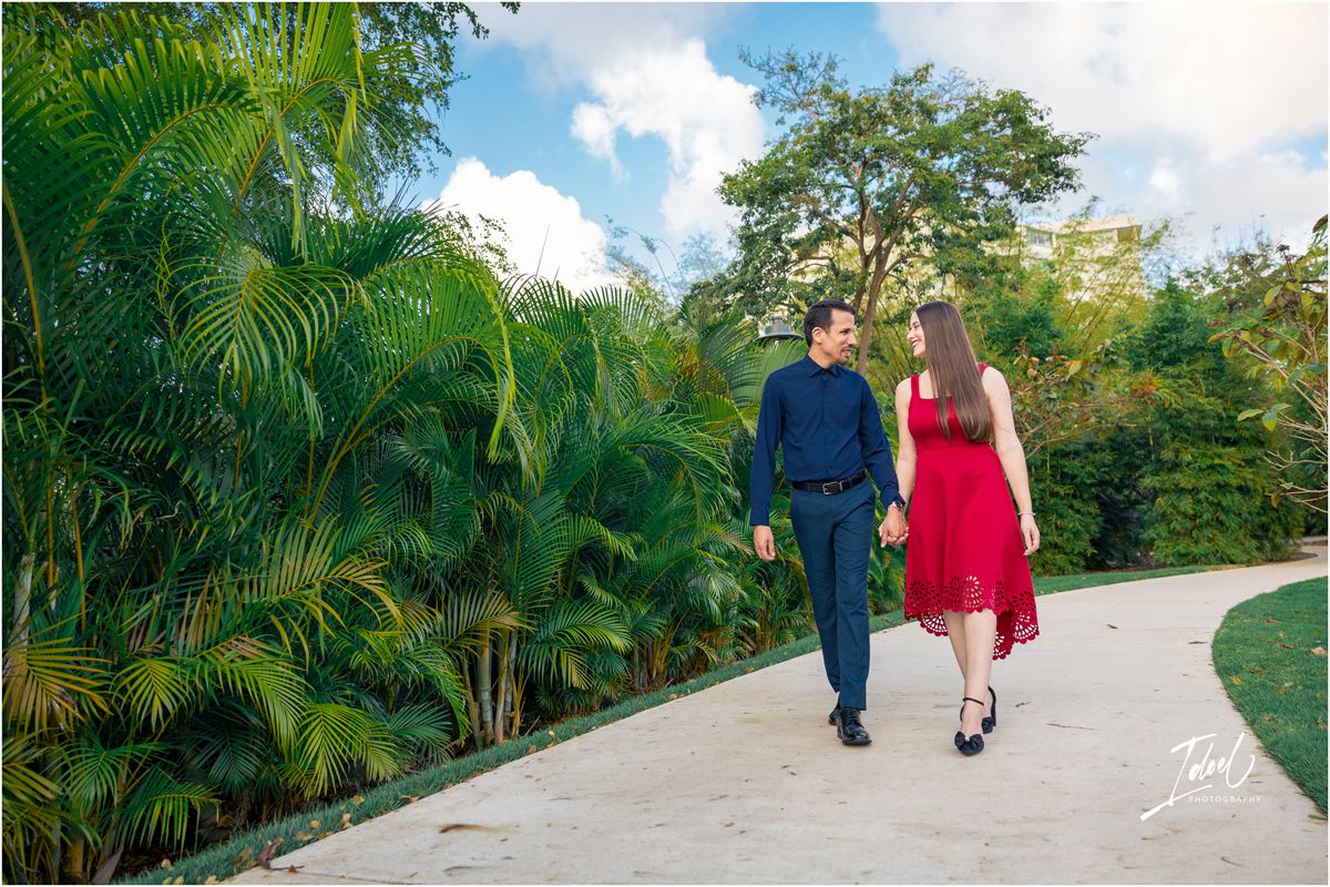 Couple holding hands walking on a garden path surrounded by lush tropical palms and trees under a blue sky.