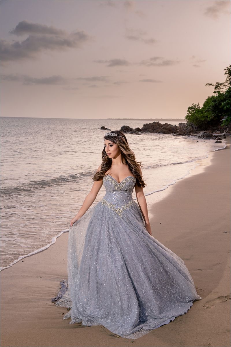 A woman in a sparkling silver ballgown twirls gracefully along a sunset beach shoreline.