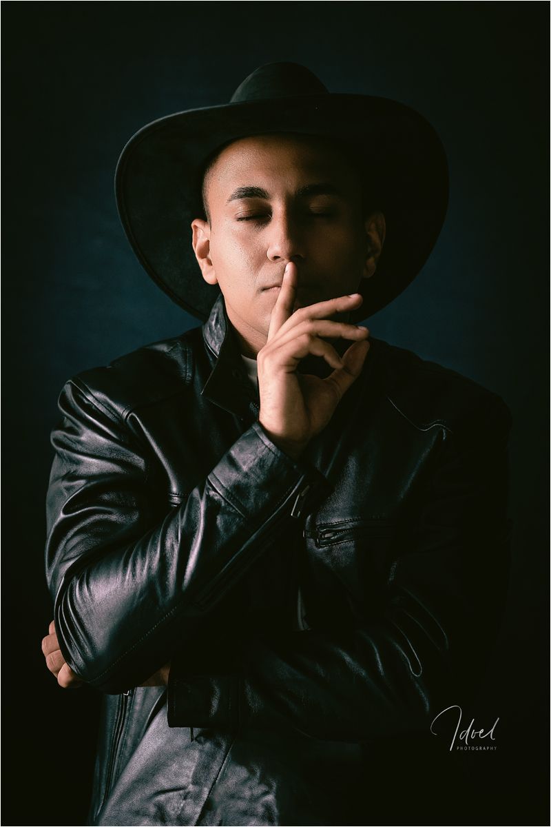Dramatic portrait of someone in a cowboy hat and leather jacket smoking against dark background.
