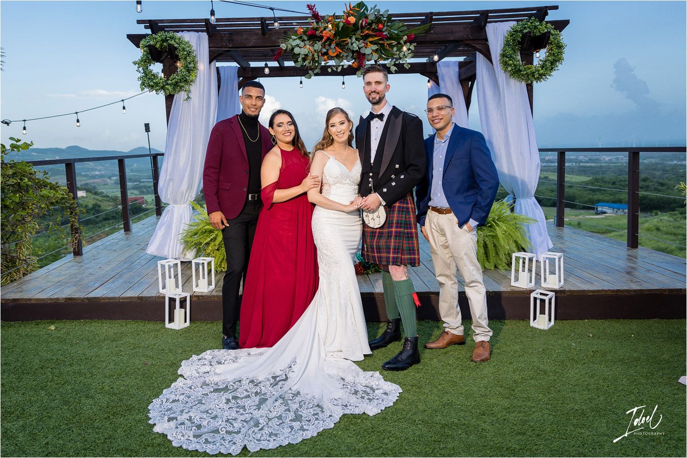 A wedding party poses under an outdoor arbor decorated with greenery and white drapery overlooking scenic mountains.