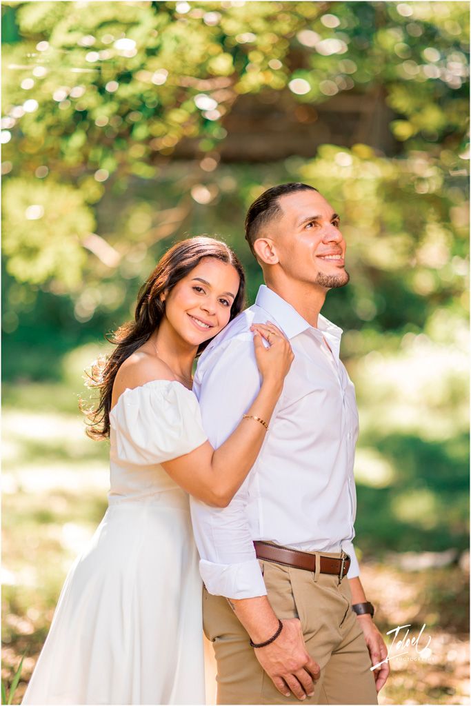 A romantic outdoor portrait of a couple embracing in white and casual attire with sunlit trees in the background.