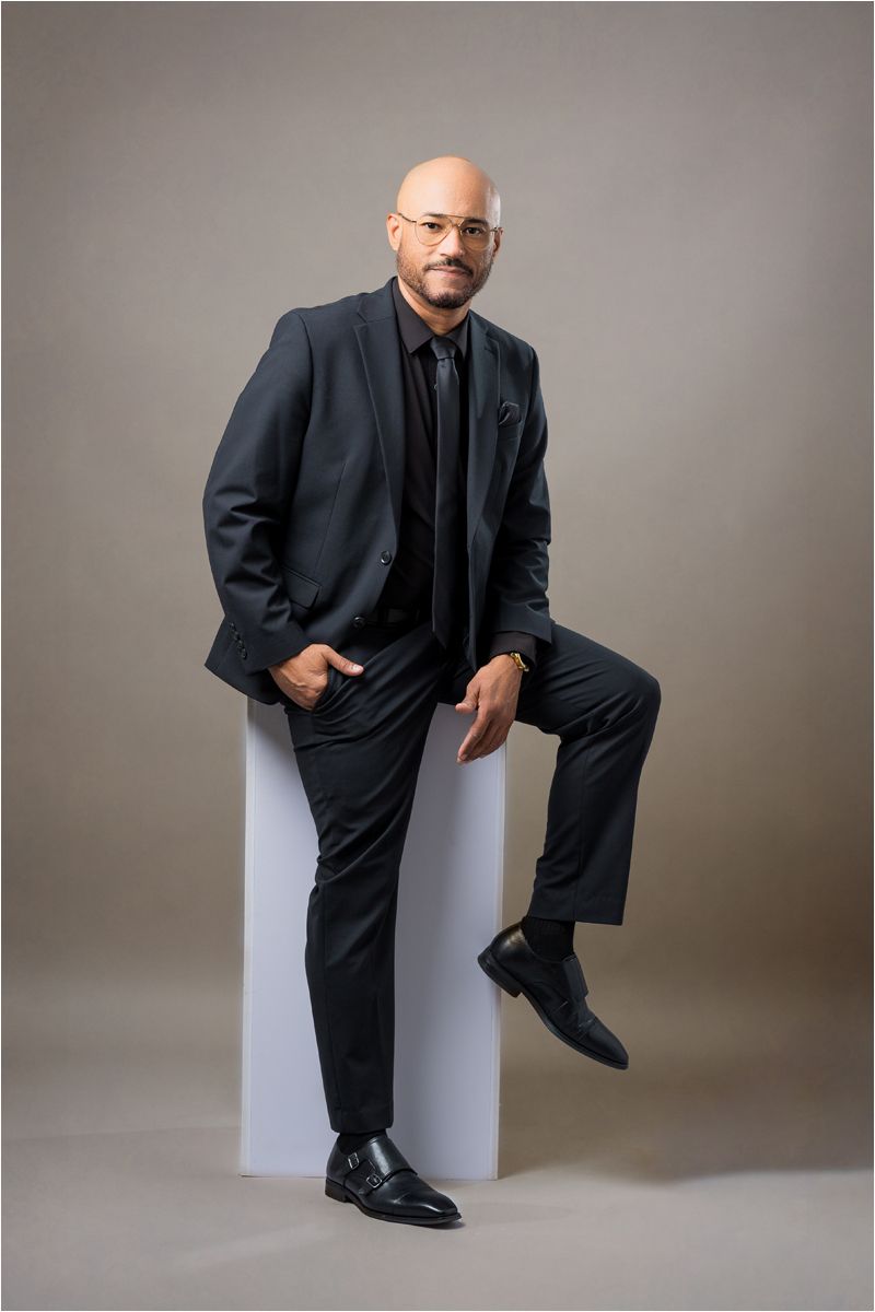 Professional studio portrait series showing a figure in a dark suit seated on a white pedestal against a neutral backdrop.