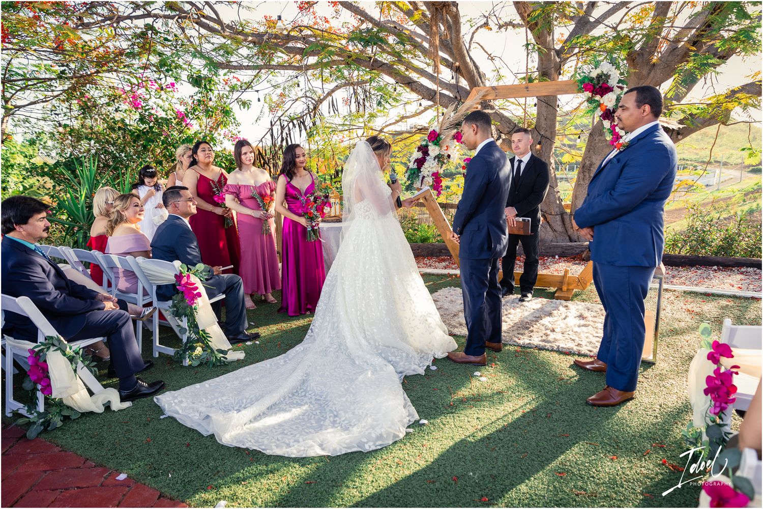 Garden wedding ceremony under flowering trees with bridal party in magenta dresses.