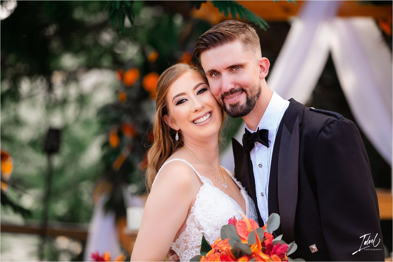 A romantic wedding portrait featuring a fitted lace gown and traditional black tuxedo with orange florals outdoors.
