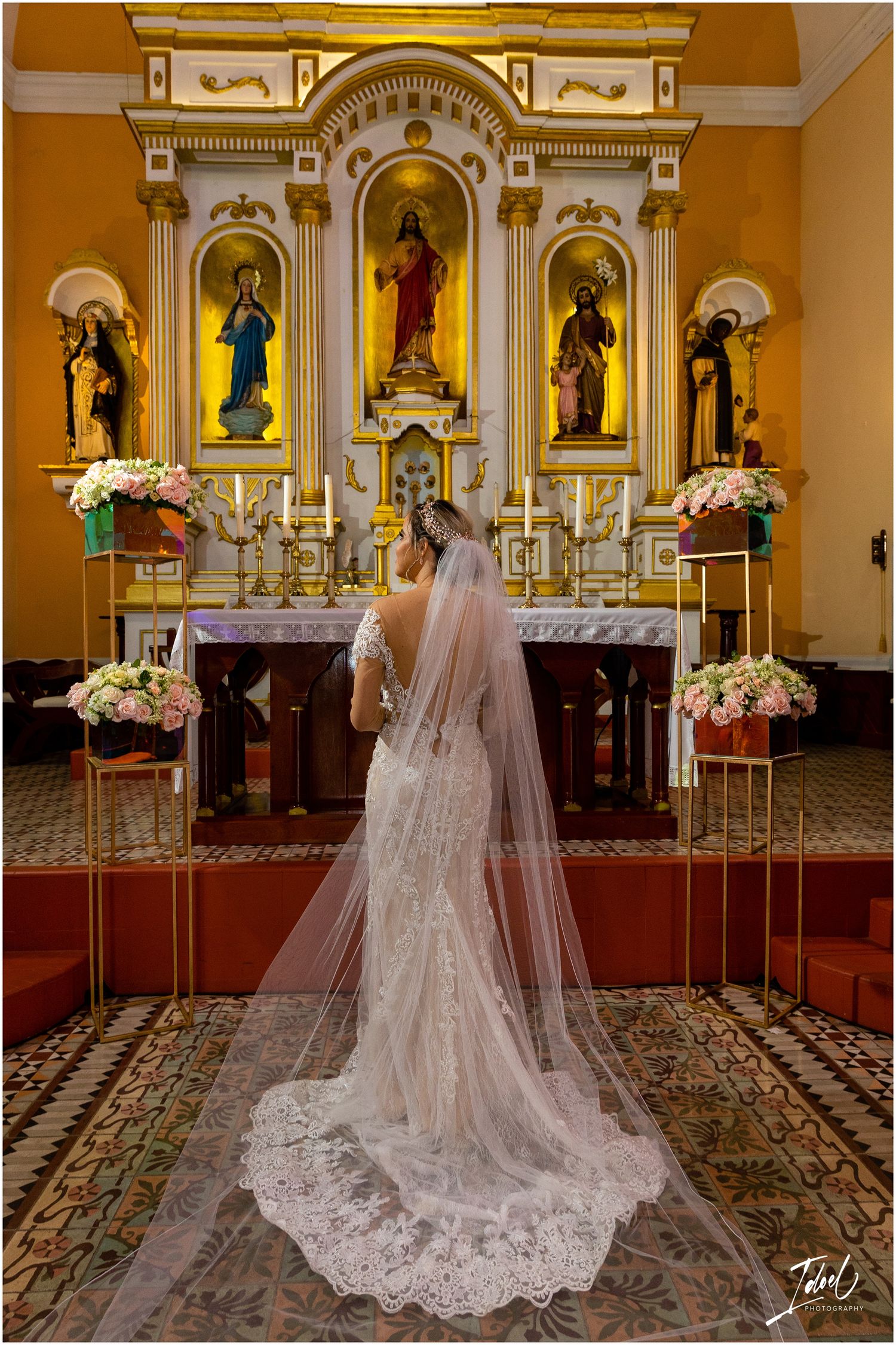 Bride in lace wedding gown with long train stands before ornate church altar with floral arrangements.