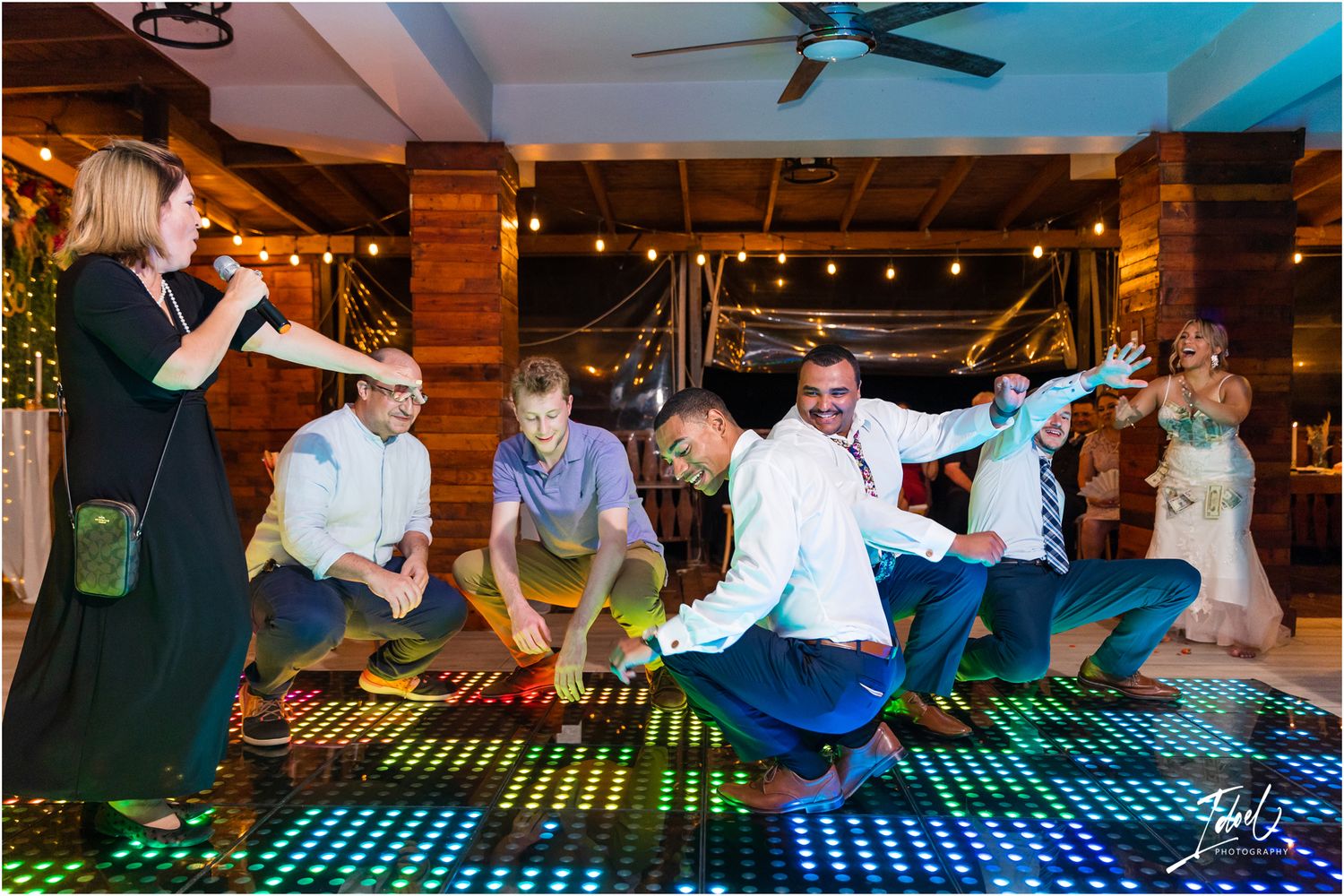 Wedding party members dancing and jumping on an illuminated dance floor during celebration.