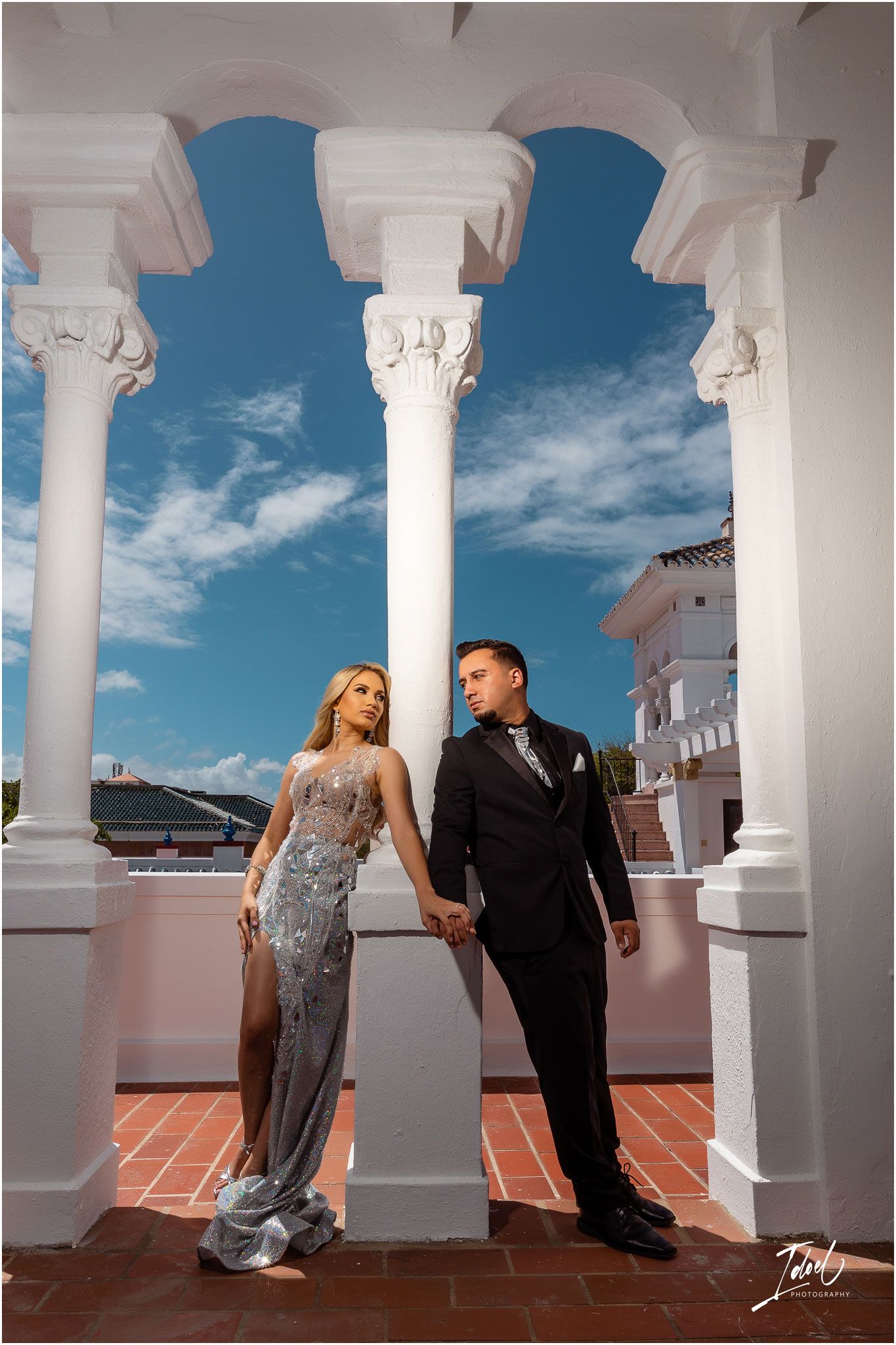 Stylish couple poses between elegant white columns against blue sky in glamorous formal attire.