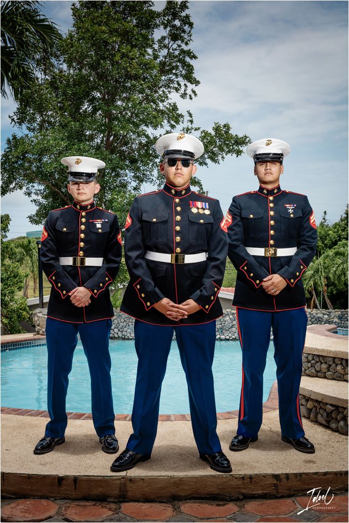 Three U.S. Marines in dress blue uniforms stand at attention by a swimming pool on a sunny day.