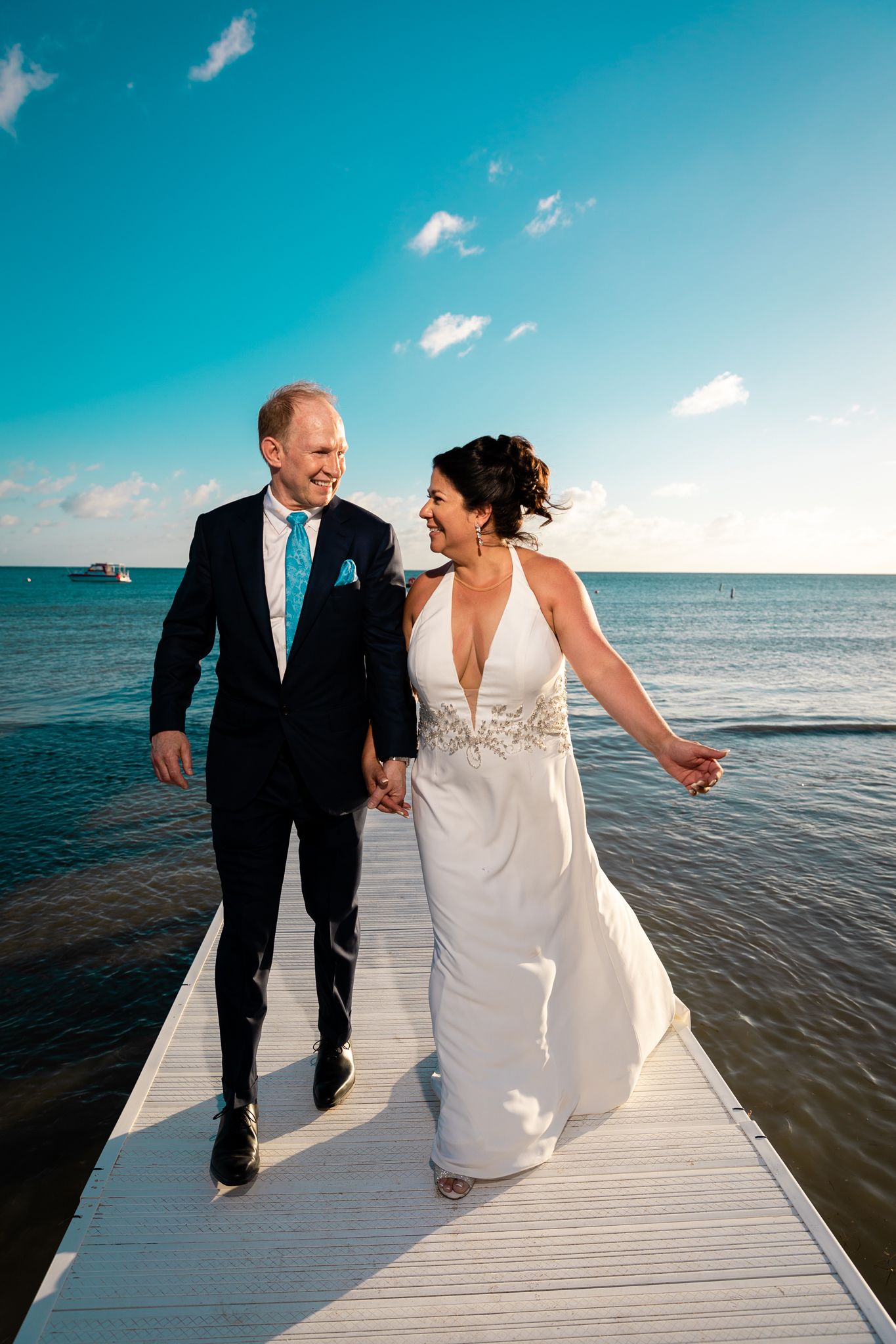 A couple in wedding attire walks and laughs together on a white boardwalk by the ocean under bright blue skies.