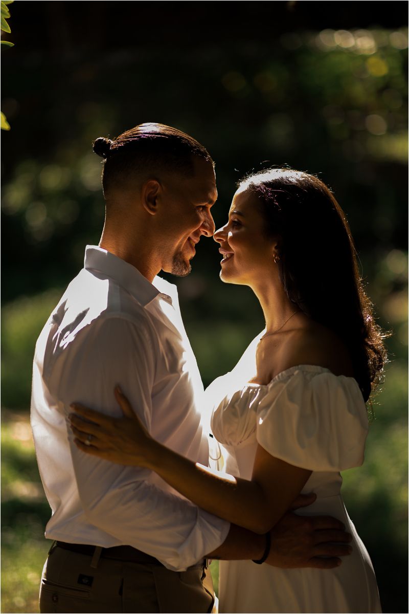 A romantic couple in white outfits embraces tenderly in a sunlit outdoor setting with natural bokeh background.