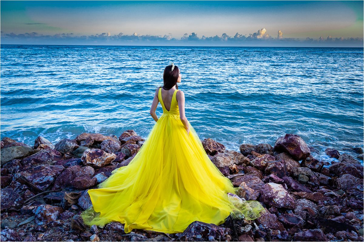 A figure in a flowing yellow gown stands gracefully against a rocky ocean shoreline at sunset.