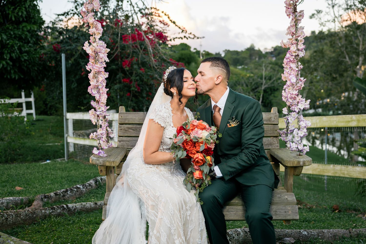 Newlyweds pose under floral archway with red bouquet in outdoor garden setting.