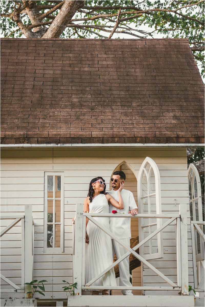 A couple in white outfits share romantic moments on a rustic porch of a white cottage with brick details.