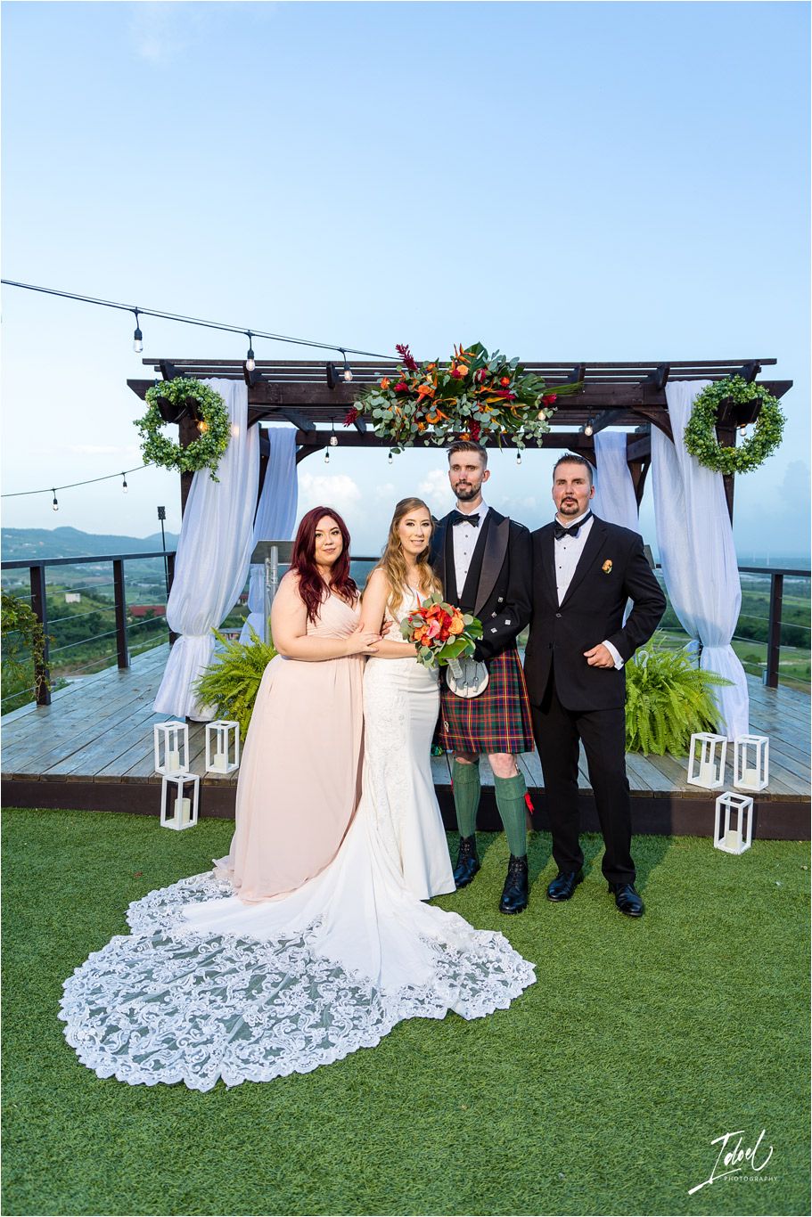 Wedding party stands against mountain view backdrop under pergola with white drapes and hanging lights.