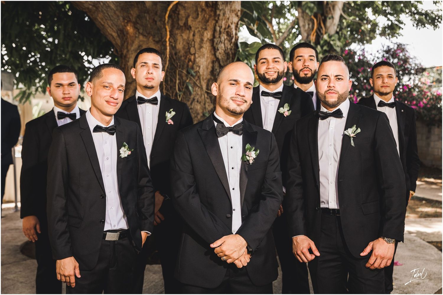 Groomsmen in classic black tuxedos pose together outdoors for formal wedding portrait.