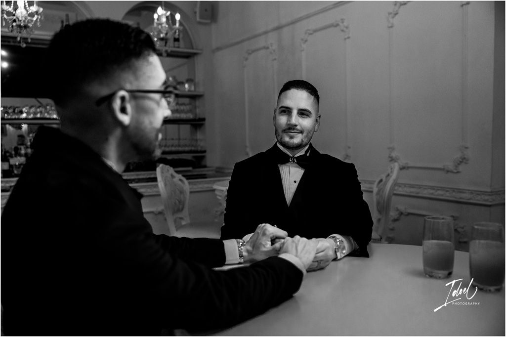 Two people in formal attire having a conversation at a table in an elegant room with chandelier lighting.