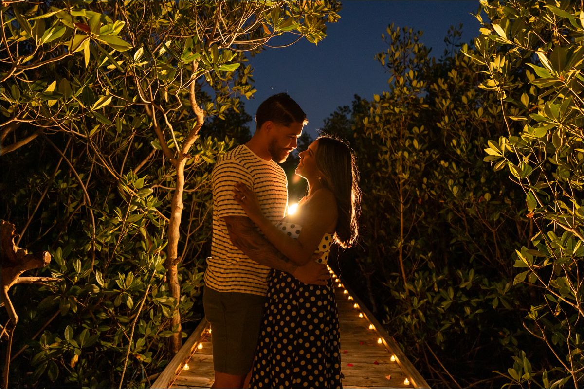 A romantic couple stands close together on an illuminated pathway surrounded by trees at night.