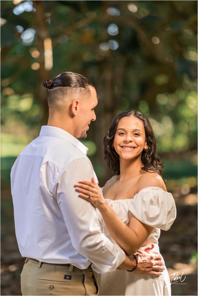 A romantic outdoor portrait of a couple embracing in formal wedding attire against a soft natural bokeh background.