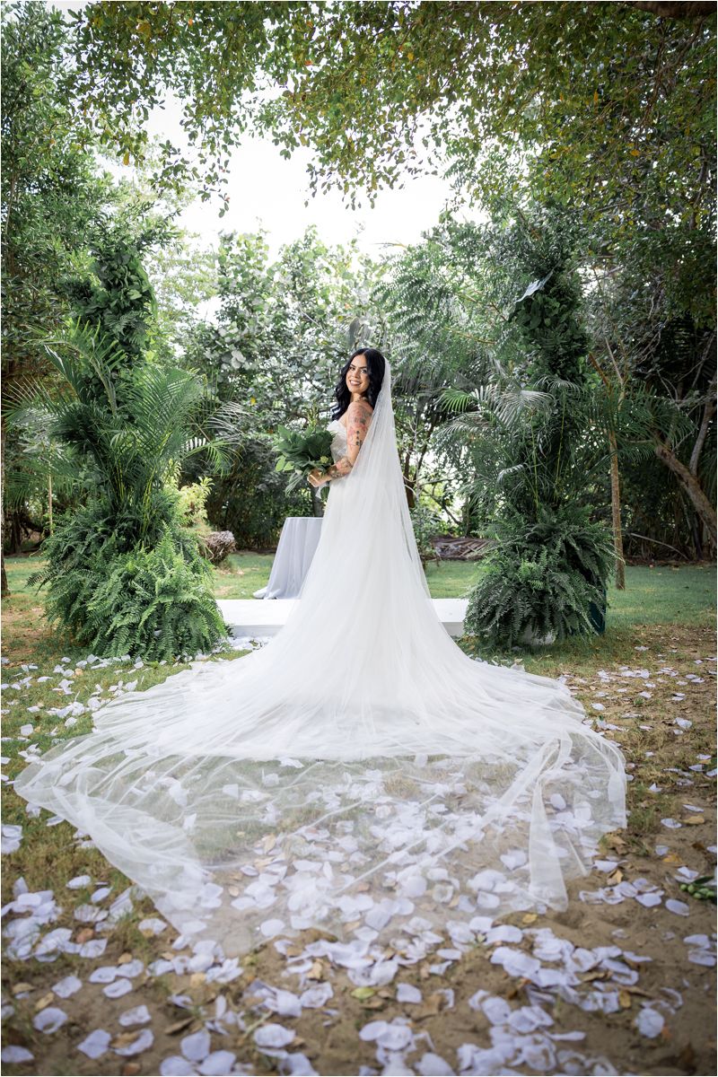 A flowing white wedding dress with cathedral-length veil trails along a garden path scattered with rose petals.