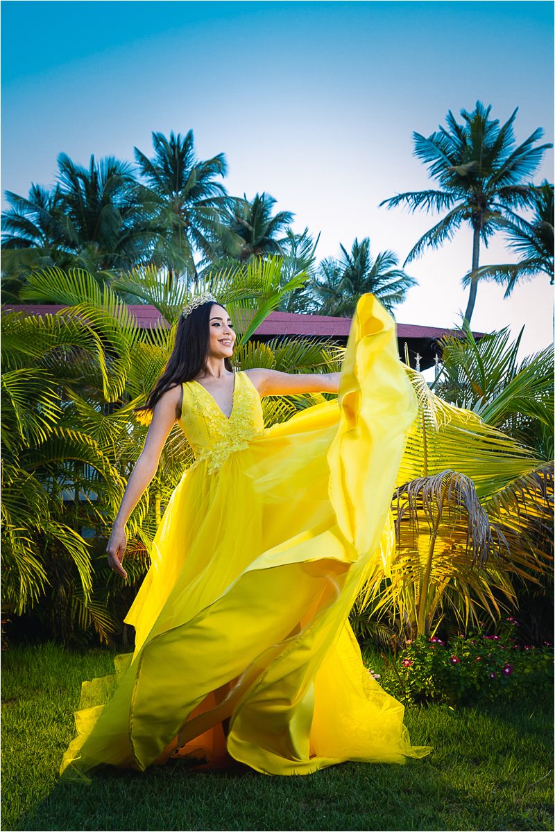 A flowing yellow gown billows dramatically against a tropical backdrop of palm trees and lush greenery.
