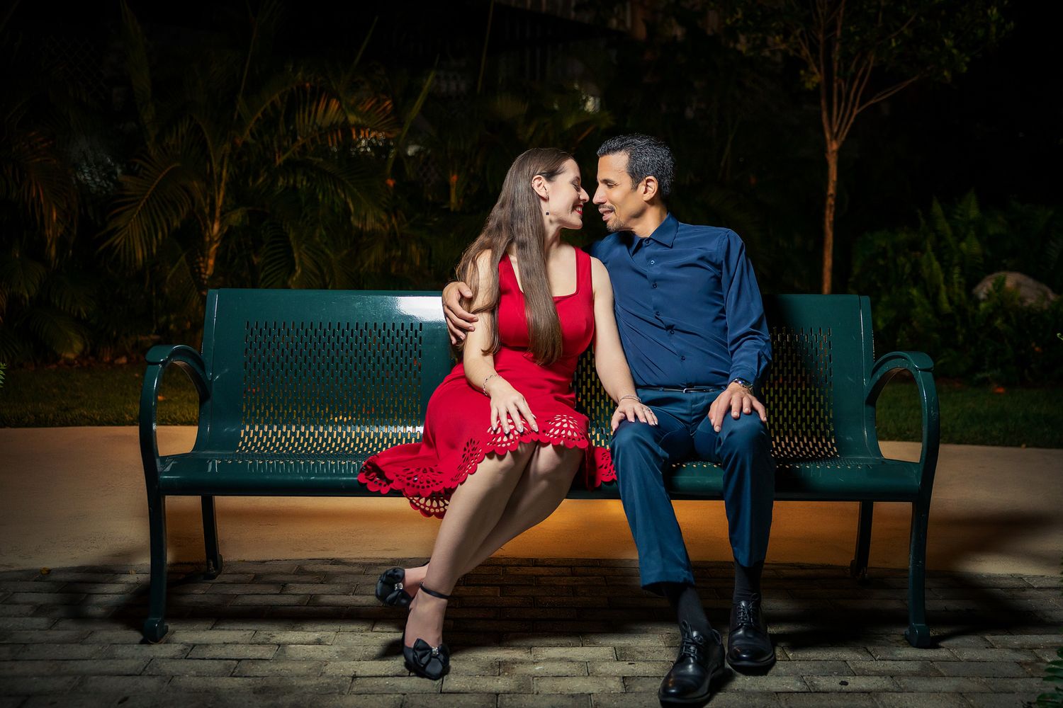 Romantic couple sitting close on a green park bench at night, woman in red dress, man in blue suit.