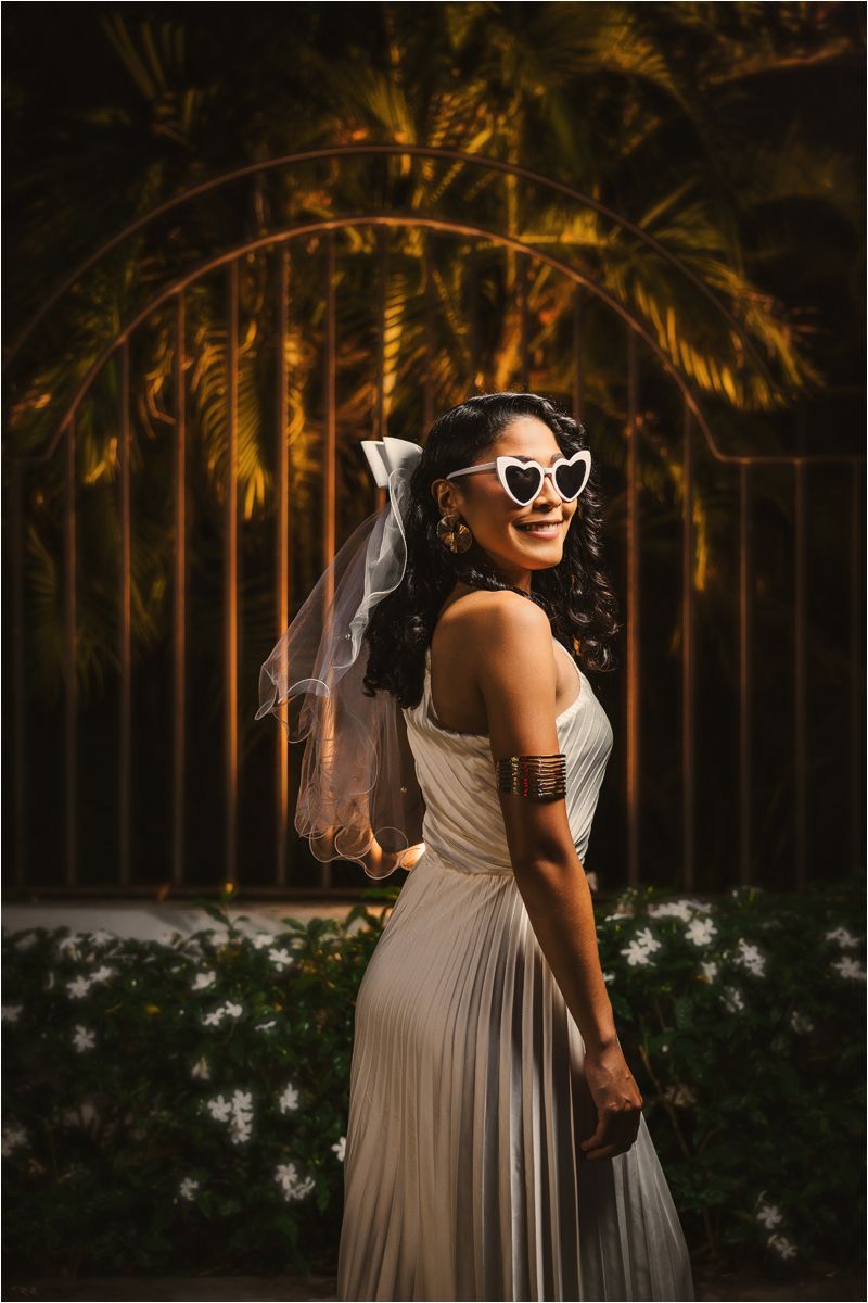 A person in a flowing beige dress and sunglasses poses in a garden with palm trees at sunset.