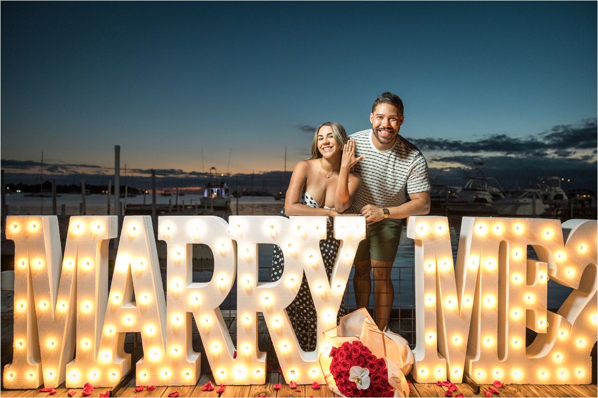 Illuminated marquee letters spell out MARRY ME against a sunset sky with red roses and people posing at dusk.