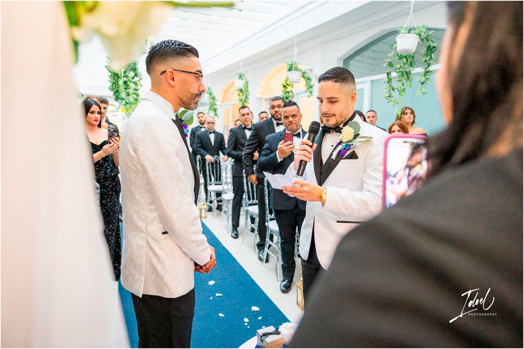 Two people in formal attire participate in a wedding ceremony on a blue carpet runner while guests look on and take photos.