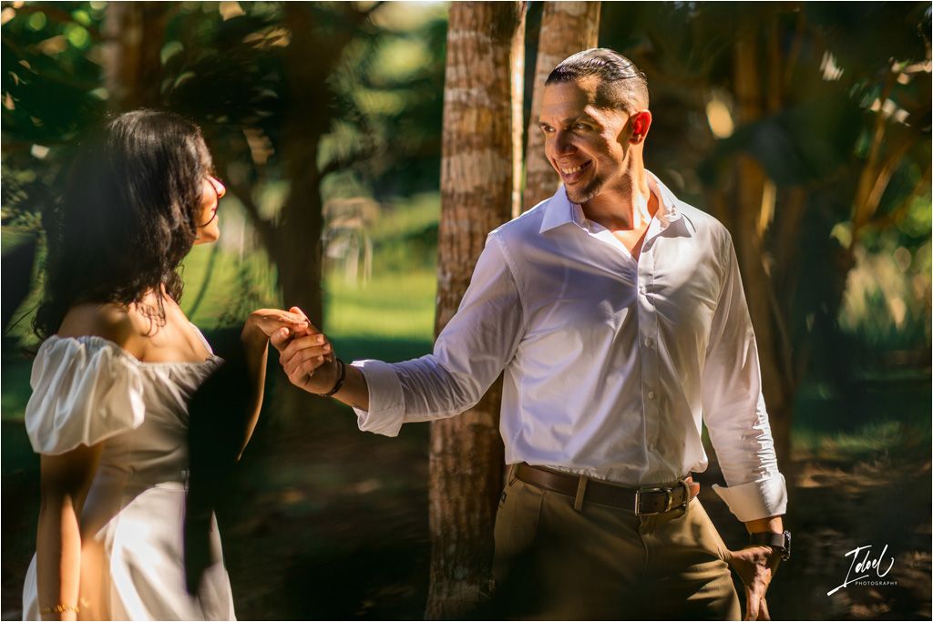 A romantic couple holding hands in a sunlit forest setting wearing white and brown attire while standing by a tree.
