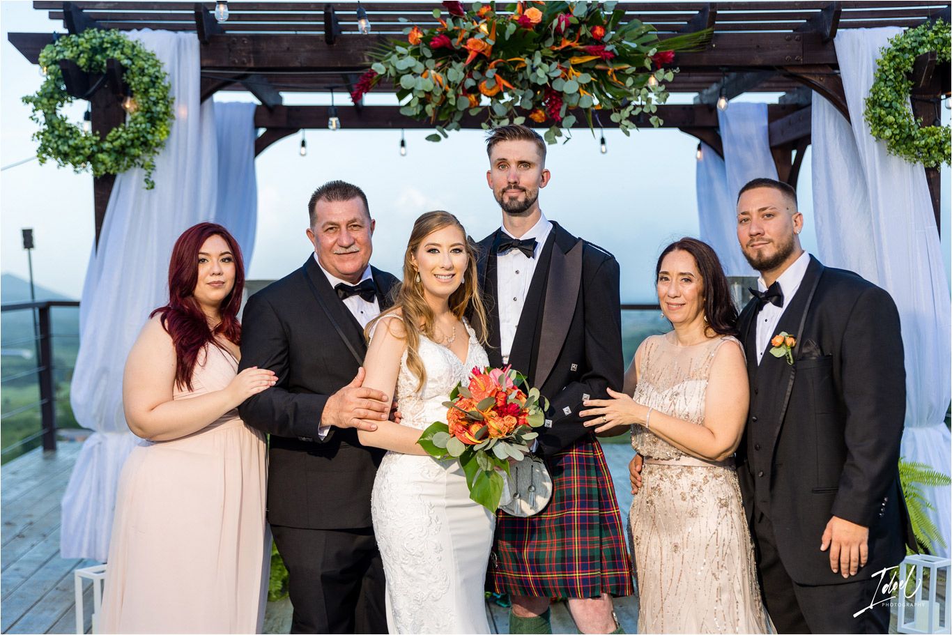 Wedding party poses under decorated outdoor pergola with white drapes and floral wreaths at scenic venue.