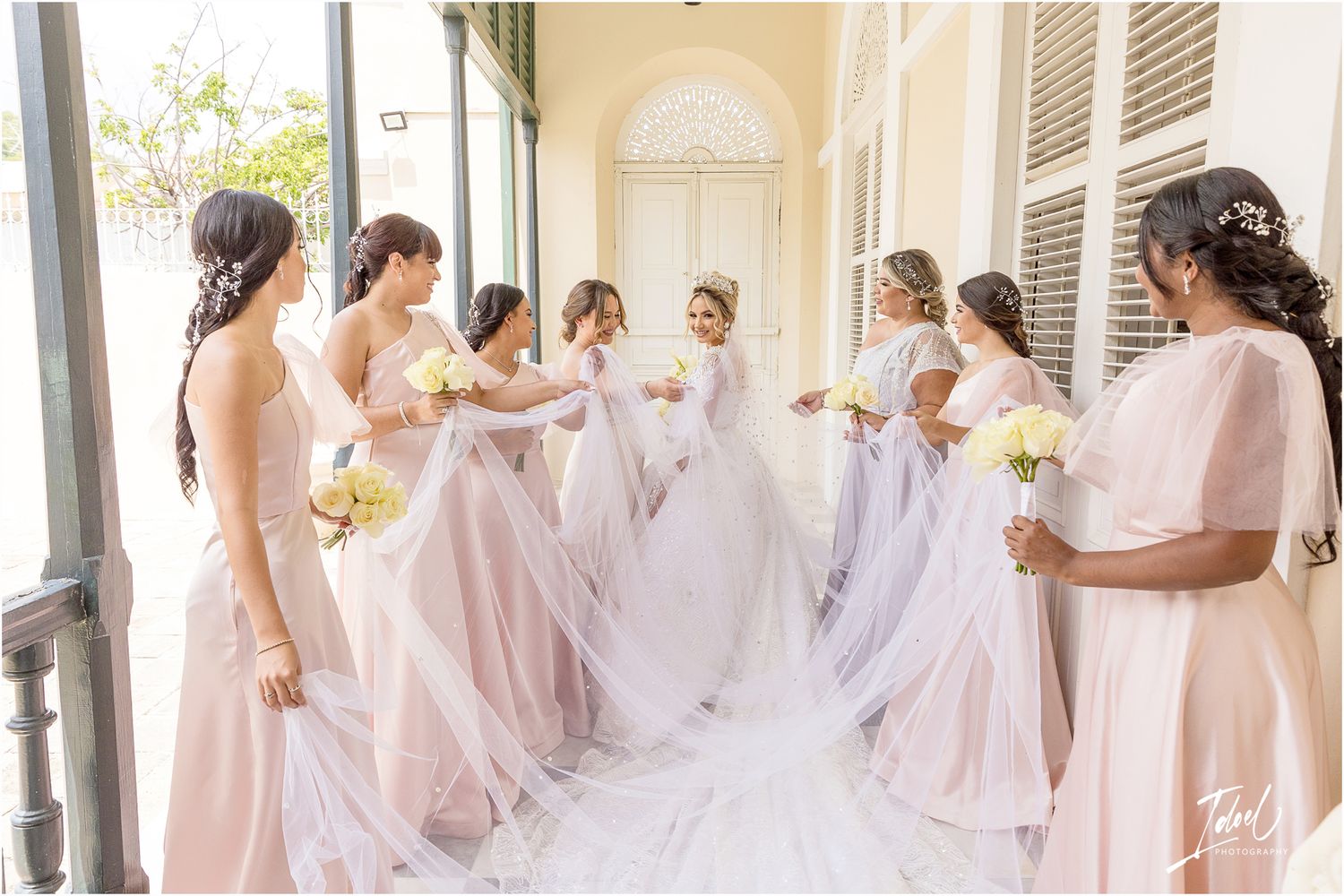 Bridesmaids in blush pink dresses gather around bride's flowing veil on covered porch.