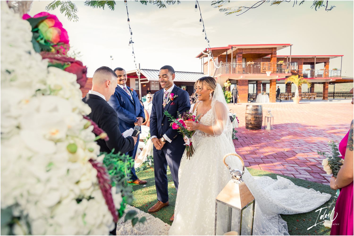 Outdoor wedding scene with vibrant floral arch and decorations against white backdrop.