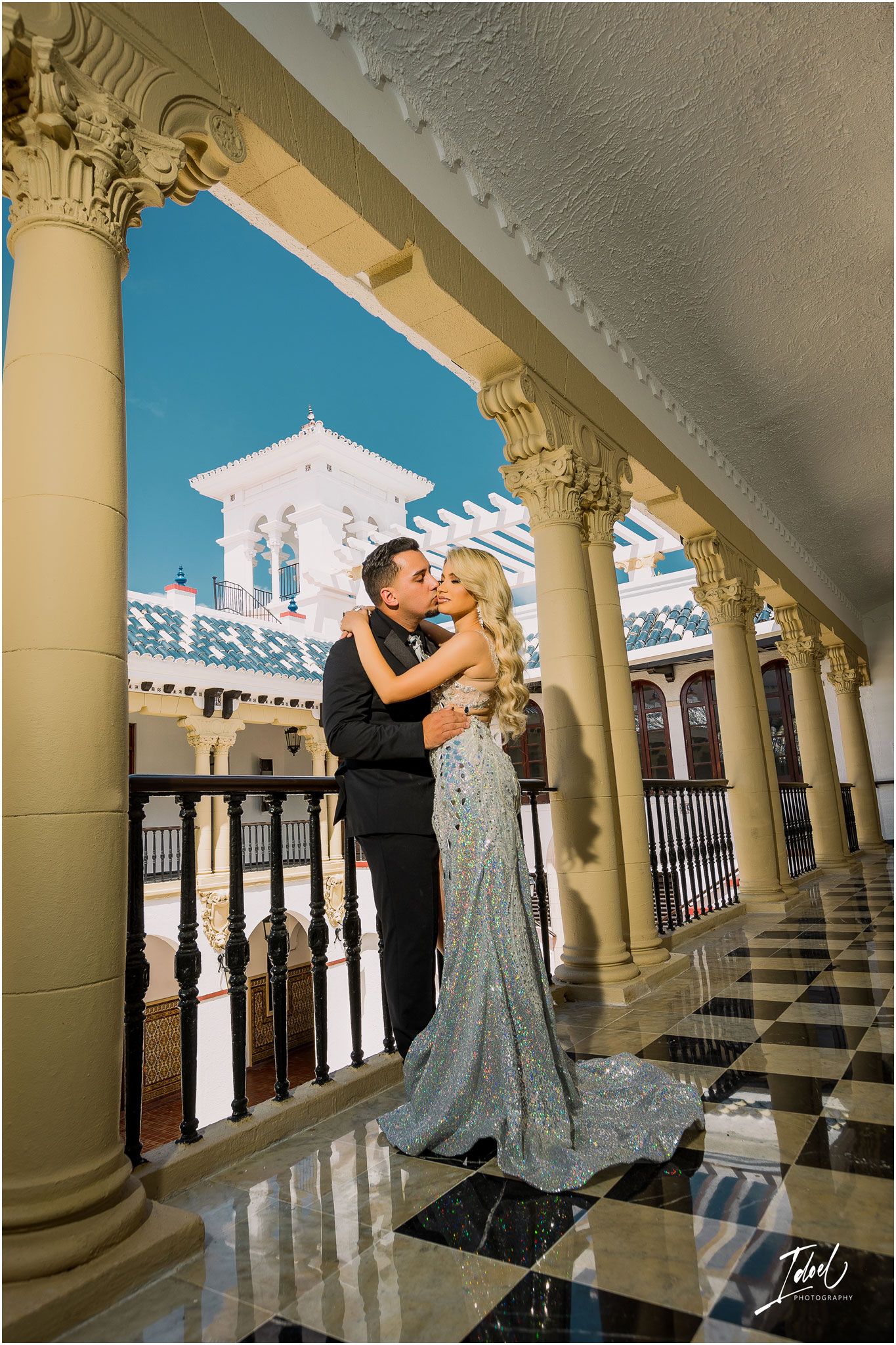 Romantic couple embracing in an ornate colonnade with Spanish architectural details and checkerboard flooring.