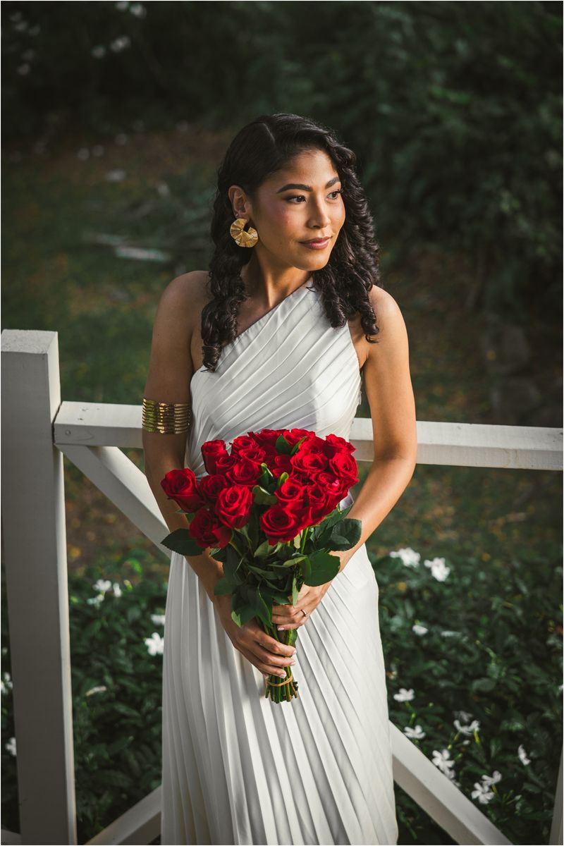 A bride in a flowing white one-shoulder gown holds a vibrant bouquet of red roses on a white porch railing.