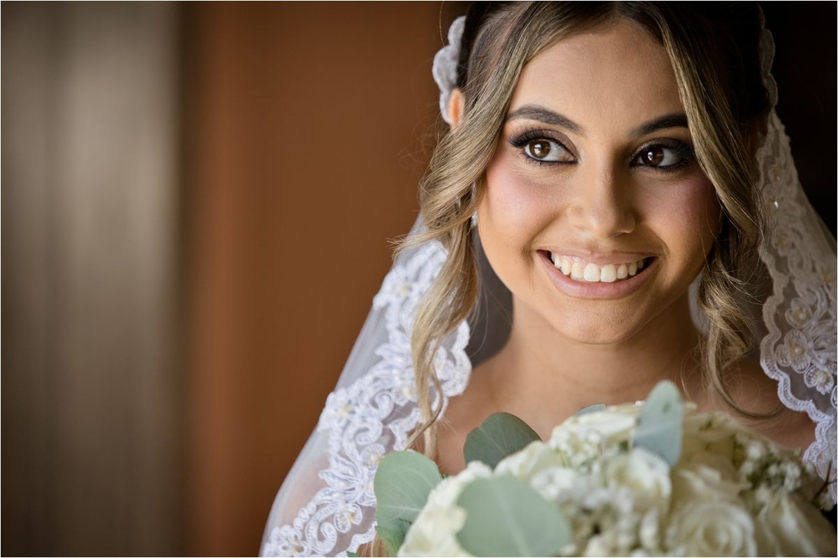 Smiling bride wearing lace veil and holding white bouquet on her wedding day.