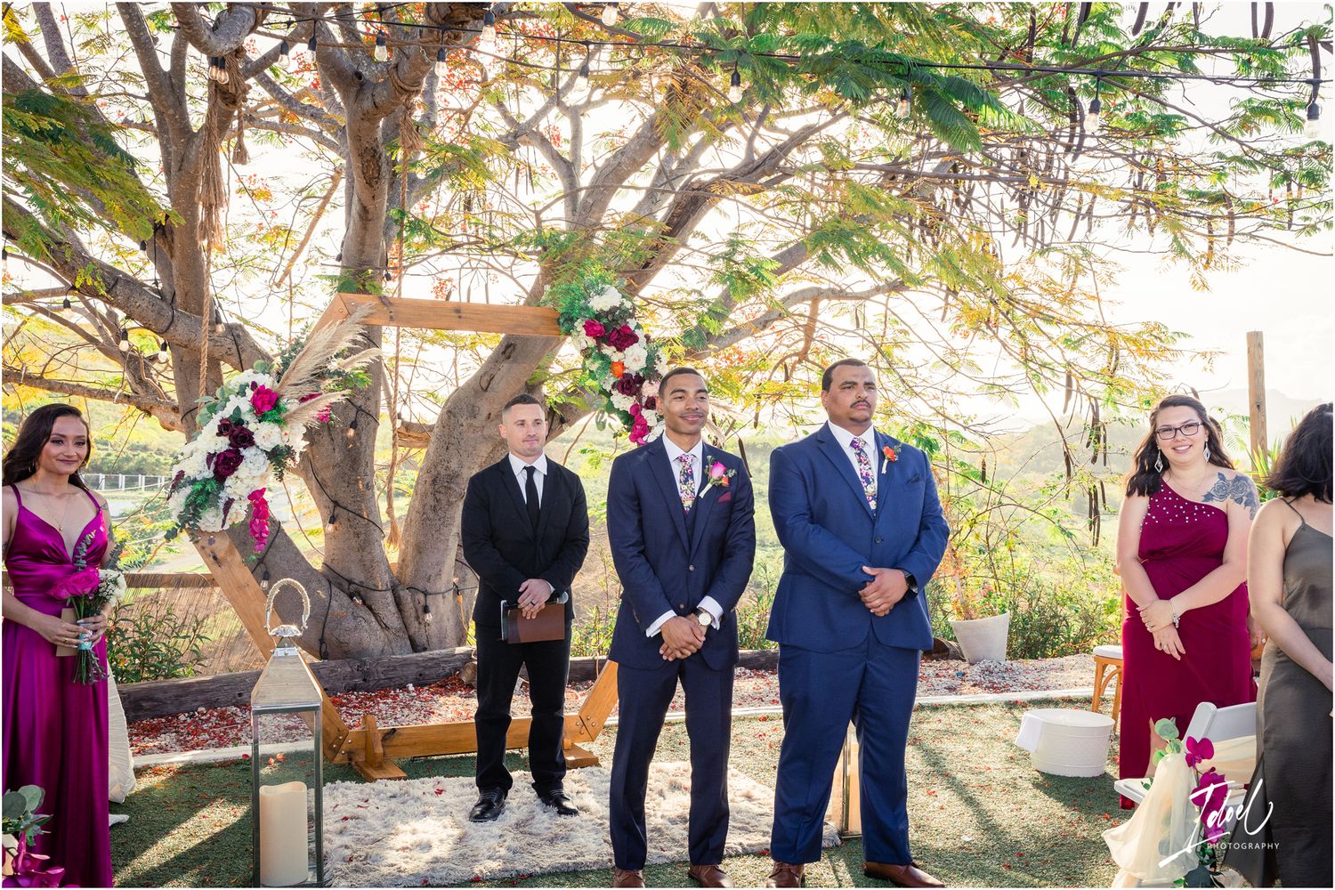 Wedding party in navy and burgundy attire standing under flowering trees during an outdoor ceremony.