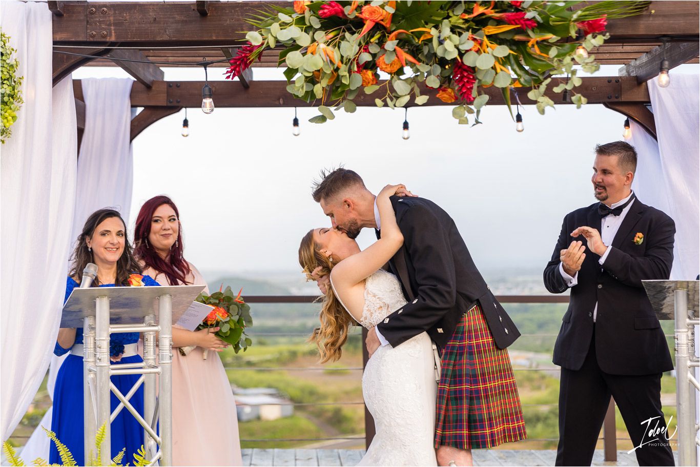 A romantic wedding kiss under a floral-adorned pergola during an outdoor ceremony.