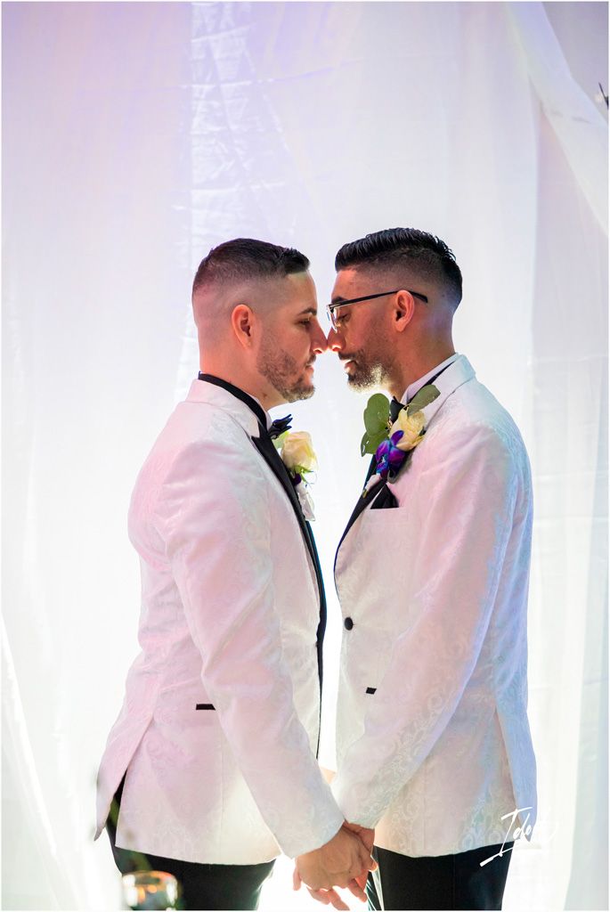 A romantic wedding moment with two people in matching white tuxedo jackets and boutonnières standing close together.