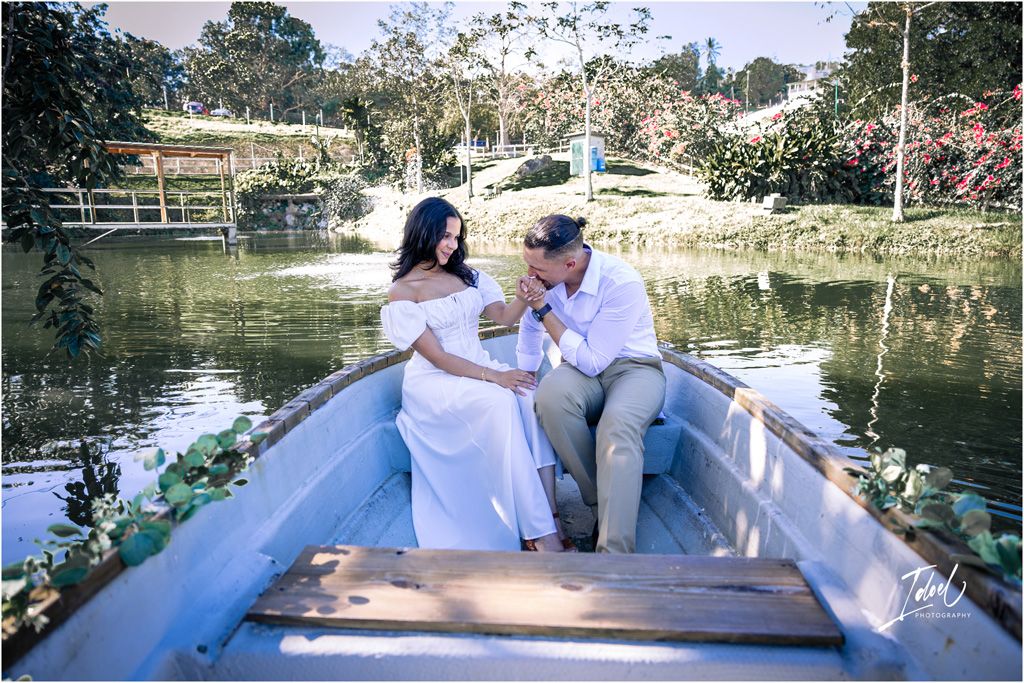 A couple sits together in a wooden rowboat on a scenic pond surrounded by trees and flowering plants at sunset.