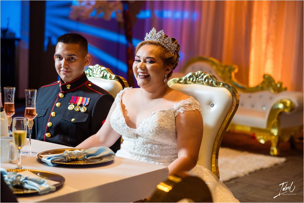 A wedding couple seated at an ornate head table with gold throne chairs during their reception.