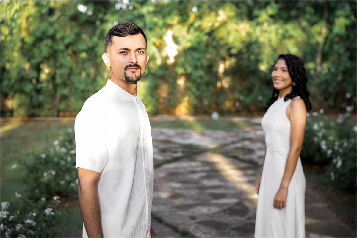 A couple in white outfits poses together during a romantic outdoor photoshoot in a sunlit wooded area.