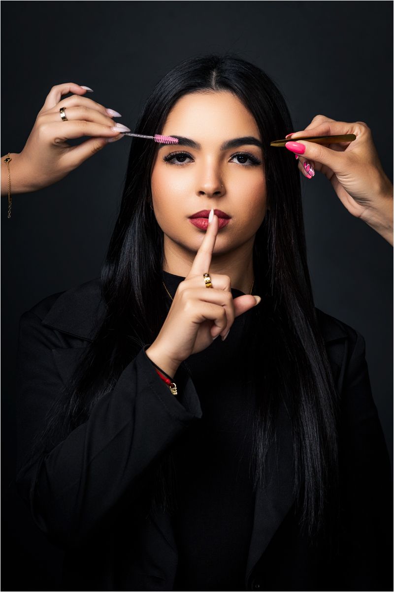 A beauty shot showing hands applying makeup with brushes while model in black makes shushing gesture against dark background.
