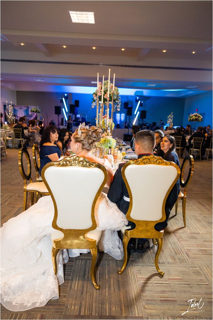 Elegant reception table setup with gold chairs and tall floral centerpieces in a ballroom with blue mood lighting.