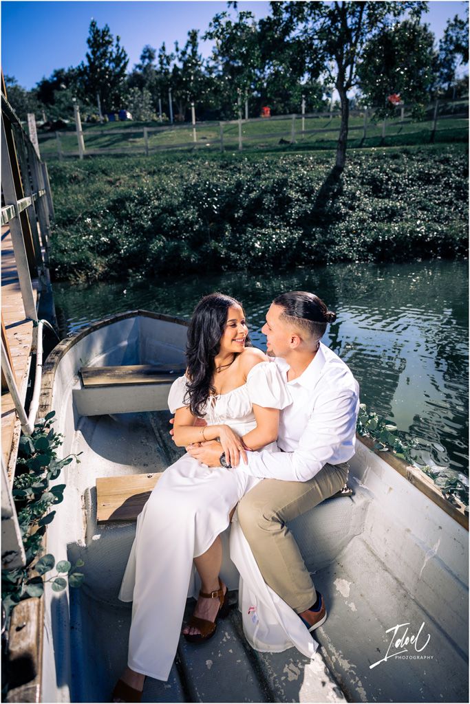 A couple dressed in white share an intimate moment while sitting together in a wooden boat on a scenic pond.