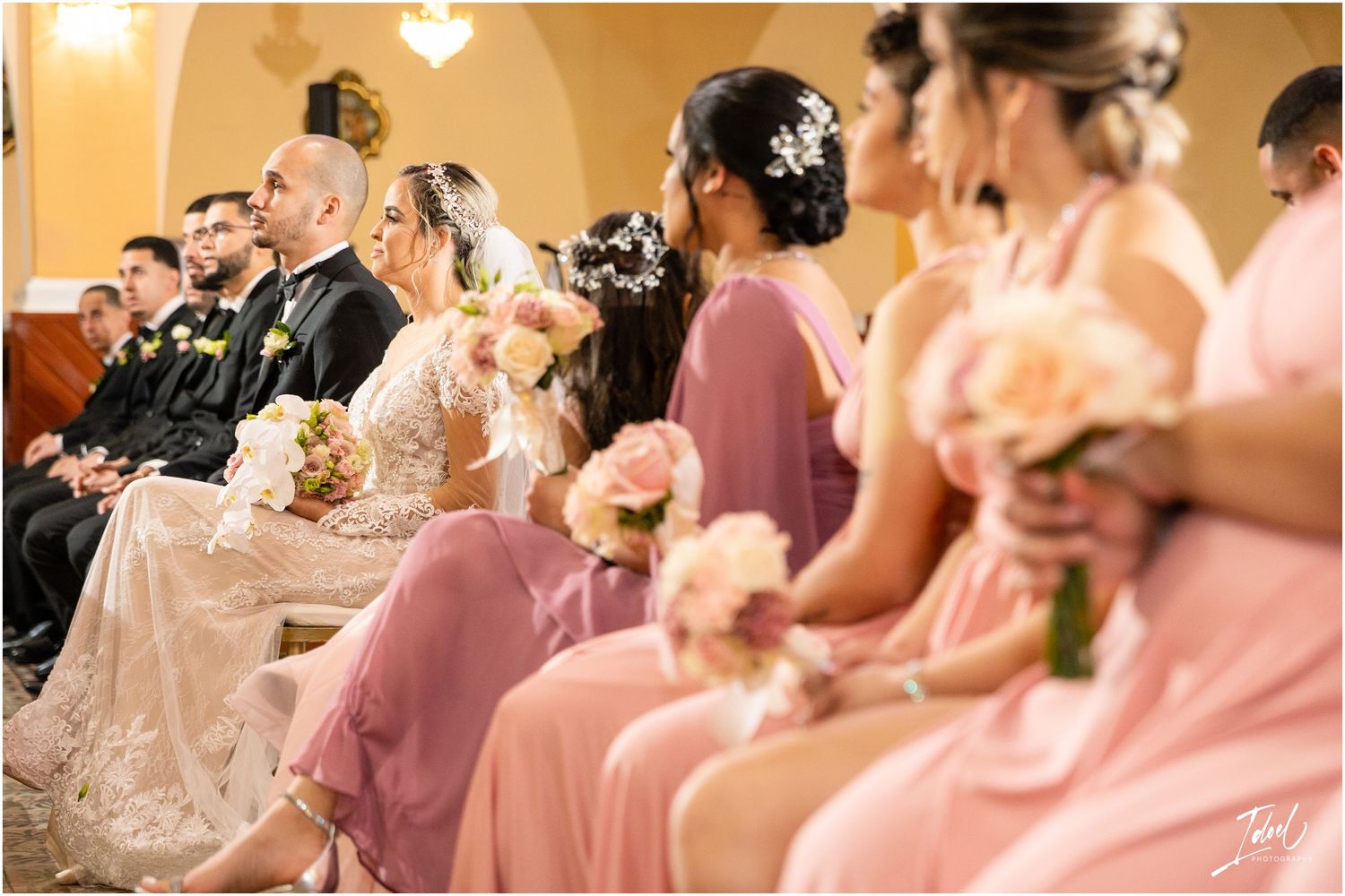 Wedding party seated in church with bridesmaids wearing pink dresses holding bouquets.