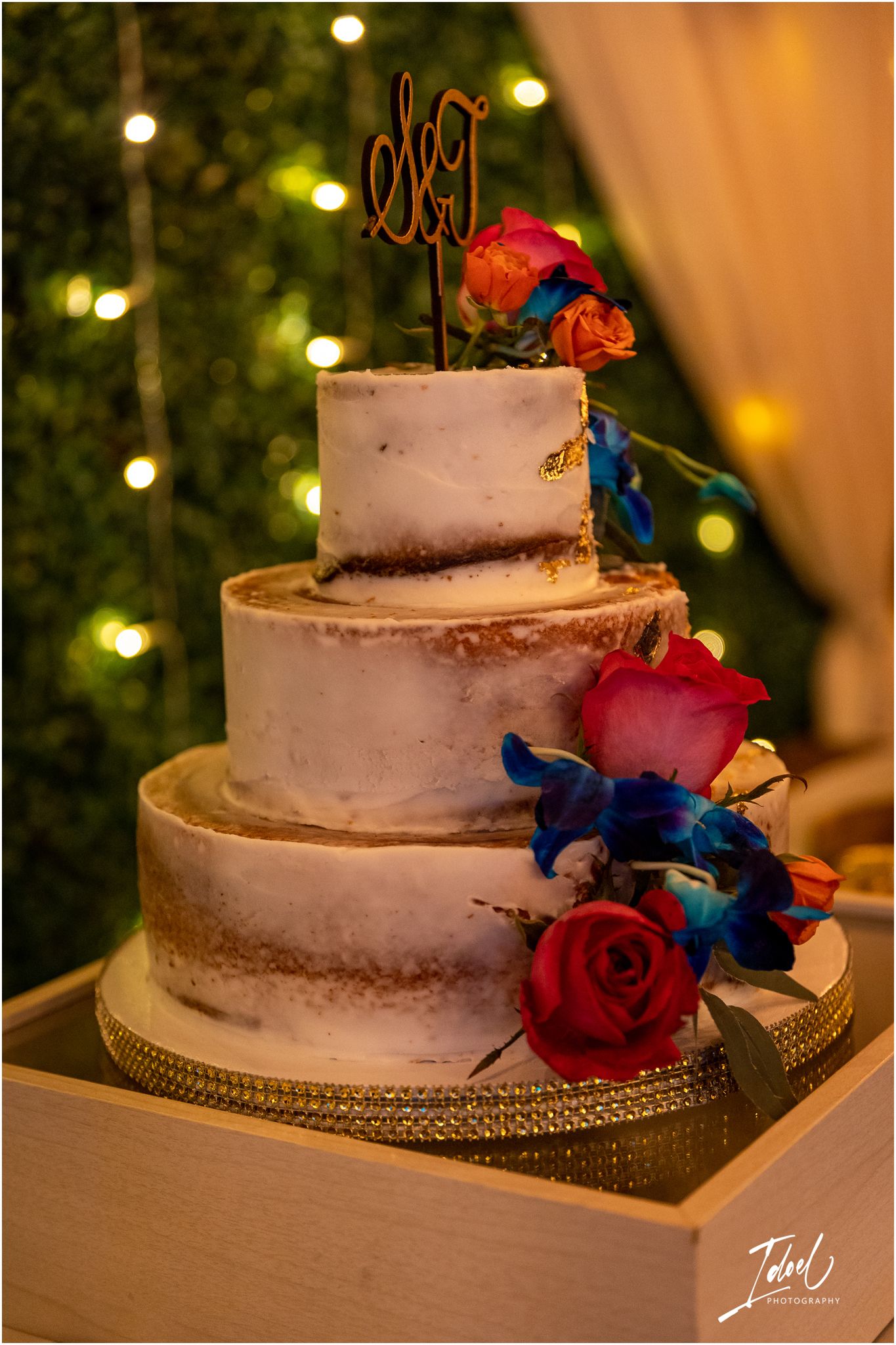 Three-tiered white wedding cake decorated with red roses and blue orchids on a gold stand.