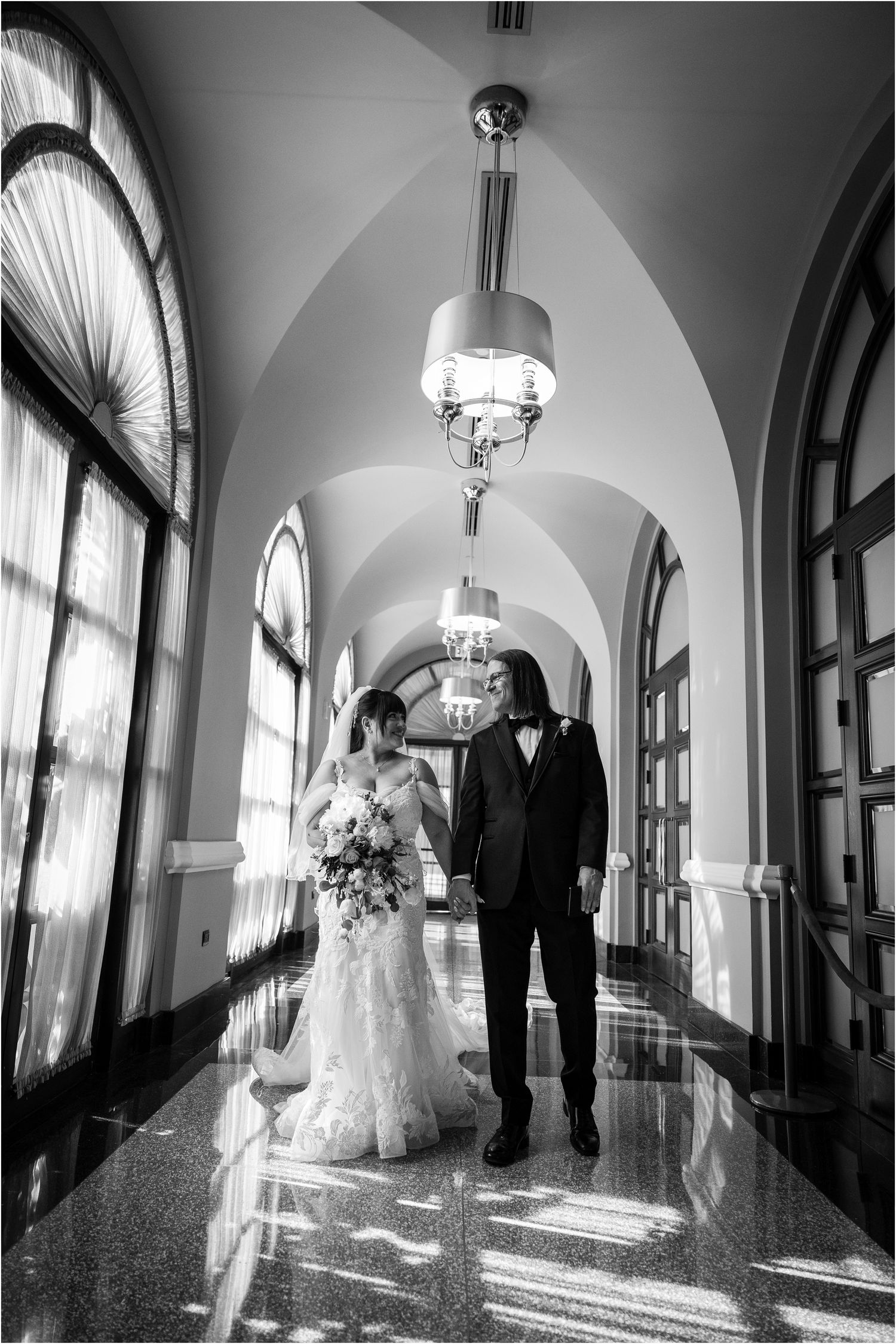 A black and white wedding photo series in an elegant hallway with arched ceilings and chandeliers.